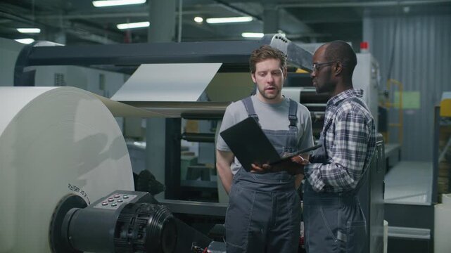 Two multiethnic industrial operators analyzing data on laptop and discussing machinery diagnostics while standing next to massive paper reel in printing factory