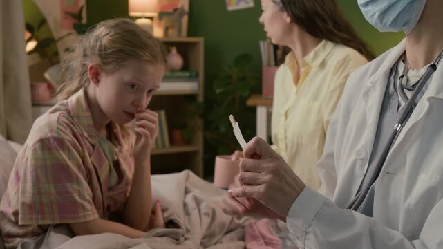 Female doctor wearing white coat and medical mask opening package and using spatula to examine throat of gen alpha girl during home visit, mother watching in background