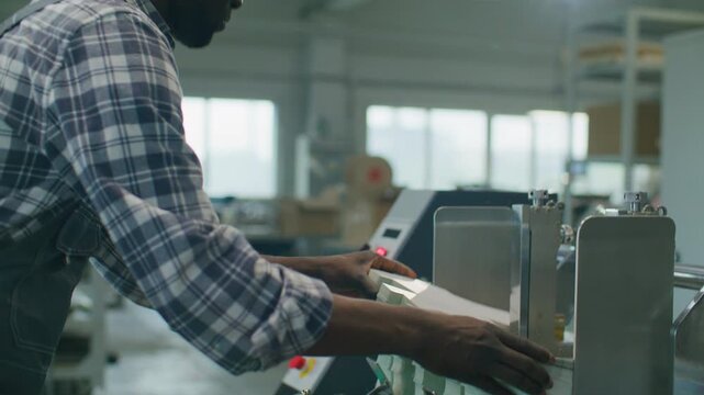 African American printing factory worker loading paper into industrial processing machine, handling printed material and monitoring automated production equipment in workshop