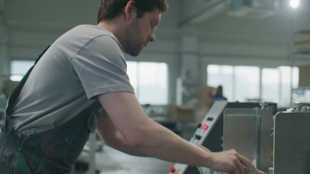 Male technician in overalls feeding paper into industrial processing machine, then pressing button on control panel while operating automated production equipment in printing factory
