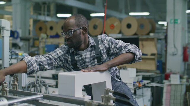 African American industrial worker adjusting mechanical clamps on automated printing press in commercial printing house, large rolls of paper visible in background