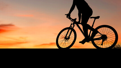 Obraz premium Silhouette of a person cycling on a mountain bike during a vibrant sunset. The sky is filled with warm orange and yellow hues, casting a dramatic glow over the landscape