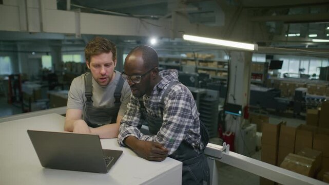 African American and Caucasian industrial engineers wearing overalls reviewing data on laptop and having professional discussion in warehouse of printing center