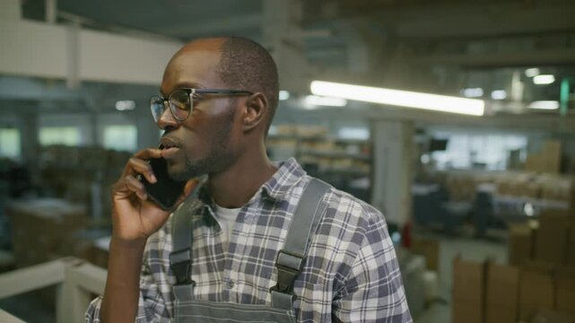African American print shop supervisor speaking on mobile phone in industrial warehouse with shipping boxes and heavy machinery visible in background