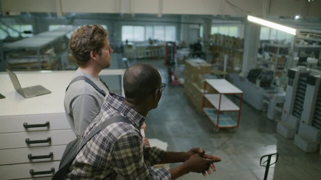 Two diverse male technicians standing on mezzanine, overlooking production floor and discussing manufacturing process in modern printing factory