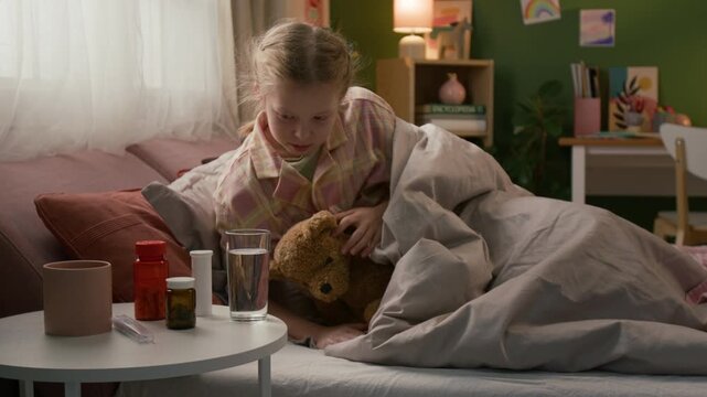 Sick gen alpha girl opening eyes and picking up glass of water from bedside table to take sip when napping with plush toy