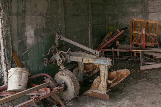 Rusted plow and old agricultural implements stored in a dark concrete shed with pallets, debris, tire, and worn machinery parts. Useful for farming, rural storage, mechanical repair, and abandoned equ