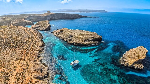 Aerial, wide, panoramic photo of the rocky coast of Comino Island in Malta, with a view of the Blue Lagoon and the yachts moored in the bay.