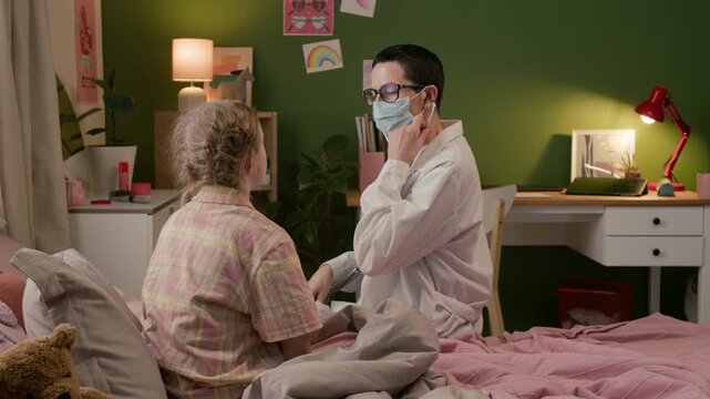 Mature female physician in lab coat inserting earpieces of stethoscope into ears and checking lungs of teenage girl during home visit