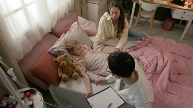 High angle view of short-haired female medical worker setting up intravenous fluid infusion for gen alpha girl and sitting down on bed beside her, mother holding daughters hand