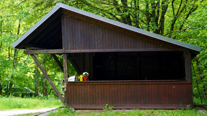 A brown wooden gazebo stands in a lush summer forest, surrounded by tall green trees and vibrant undergrowth. The rustic structure features a classic design with open sides, providing a cozy shelter a © Василий Канин