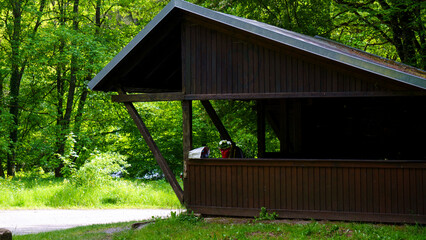 A brown wooden gazebo stands in a lush summer forest, surrounded by tall green trees and vibrant undergrowth. The rustic structure features a classic design with open sides, providing a cozy shelter a © Василий Канин