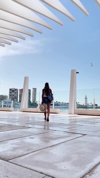 woman walks toward a lighthouse at the waterfront of the city with modern architecture and buildings under blue sky in muelle uno malaga spain