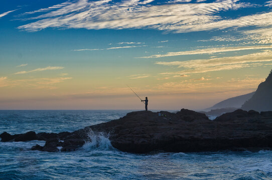 A solitary angler stands silhouetted against the dawn sky, holding a fishing rod while waves crash against the dark, rugged shorelineon the south coast of Victoria, Australia