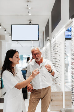 Professional female optometrist assisting a senior male customer in choosing stylish eyeglass frames at a modern optical boutique, focusing on personalized vision care and customer service.
