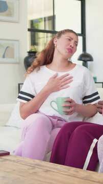 Vertical video: Smiling woman white tee taking hand as friend talking on couch with green pink mugs