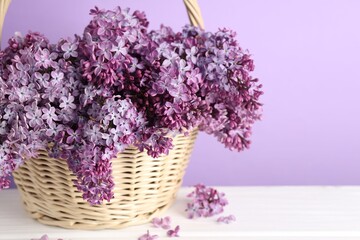 Fototapeta premium Tender lilac flowers in wicker basket on white wooden table against violet background, closeup. Space for text