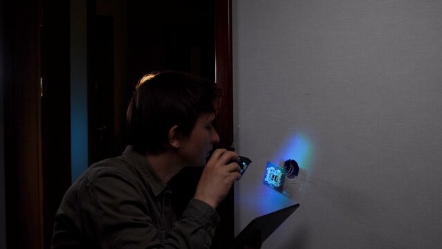 Young man holding a signal detector, searching an electrical outlet on a wall for hidden cameras or listening devices, focusing on privacy and security threats