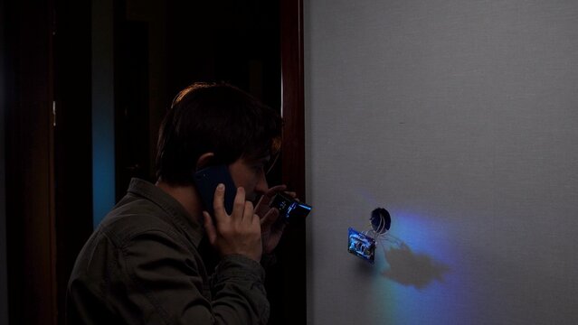 Man holding flashlight and phone, inspecting a broken electrical outlet on a dark wall, seeking guidance for urgent home electrical repair or emergency assistance and safety