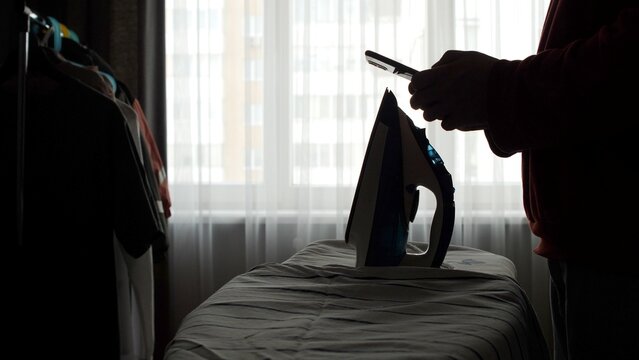 Dark silhouette by an ironing board, phone in hand, distracted from chores as soft window light casts shadowsmodern domestic life, multitasking and screen time interrupting home duties