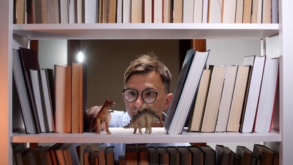 Man in eyeglasses looking curiously at dinosaur toys, including a stegosaurus and velociraptor, placed among books on a white bookshelf, reflecting childhood imagination and learning © Aliaksandr
