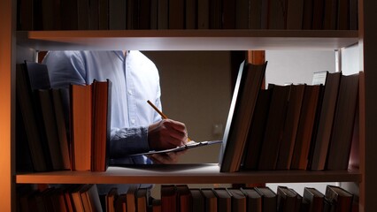 Man standing between bookshelves in a library or archive, focusing on writing notes with a pencil on a clipboard, symbolizing diligent research and deep historic study © Aliaksandr