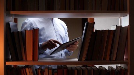 Man holding and interacting with a digital tablet while standing between bookshelves filled with traditional books, representing modern technology blending with classic learning environments © Aliaksandr