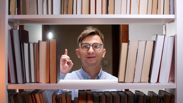 Young man wearing eyeglasses standing between bookshelves, having an exciting eureka moment with an index finger raised and a happy, surprised expression