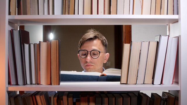 Man wearing glasses deeply focused on reading a book, partially obscured by bookshelves, representing education, wisdom, and research in a personal study or library setting