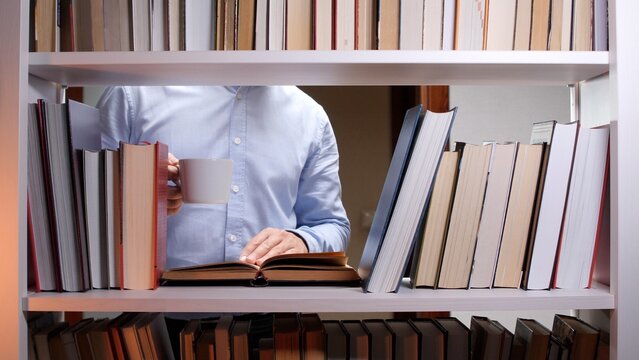 Man standing between bookshelves, reading a book and holding a hot beverage, representing education, knowledge, learning, and self improvement in a comfortable setting