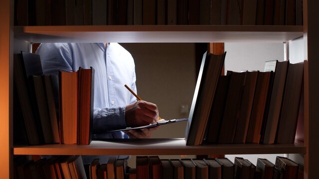 Man standing between bookshelves in a library or archive, focusing on writing notes with a pencil on a clipboard, symbolizing diligent research and deep historic study