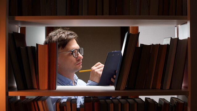 Young man wearing eyeglasses standing behind library shelves, carefully writing on a clipboard with a pencil, focusing on research and knowledge acquisition in a academic setting