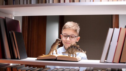 Young boy wearing glasses smiling while reading a book with toy dinosaurs on its pages, sparking imagination and a love for learning in a library setting © Aliaksandr