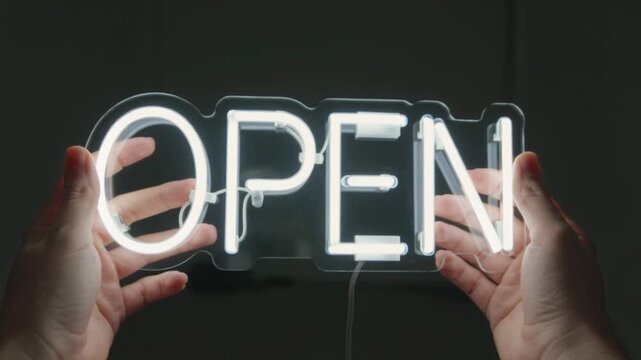 Manos sosteniendo un letrero luminoso de ne&oacute;n blanco con la palabra OPEN sobre fondo oscuro