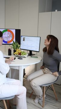 Vertical screen young woman worries about excess belly fat during nutrition consultation while dietologist encourages healthier eating habits and balanced food choices with fresh fruit