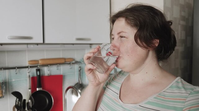 Portrait of smiling, candid plus size woman drinking water in imperfect home kitchen. Drinking clean water and maintaining hydration regimen is important for those who are overweight.