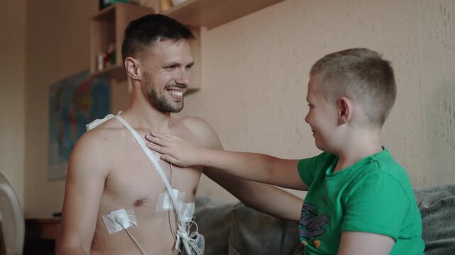 Son touches sensor of Holter monitors on fathers body and gives thumbs-up. Family responds positively to cardiac monitoring and health checkups after 30 years.