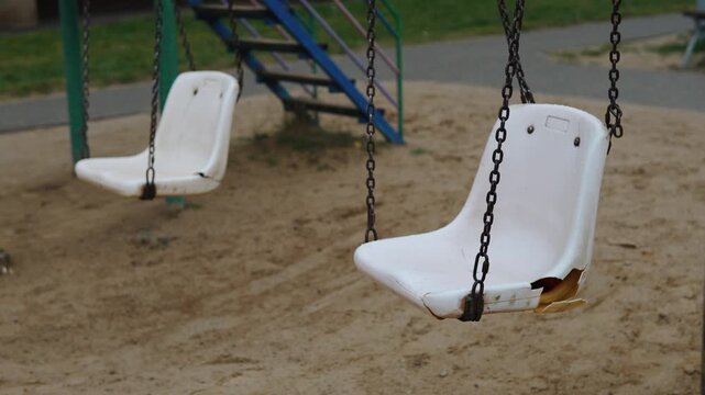 Broken, empty plastic swing seat in gloomy setting. Old, cracked and holed swing swaying on chains outside on playground. Childhood sadness and tragedy.