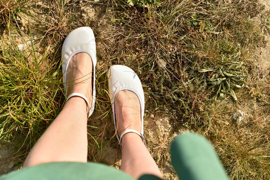 Top down view of a woman legs and feet standing on dry grassy ground wearing white barefoot ballet flats with ankle straps.