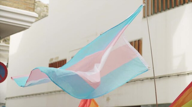 Transgender flag on flagpole moving during pride parade with LGBT flags in city. June pride parade in European Spanish city without recognizable faces.