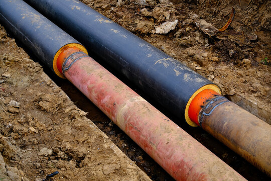 Installation of pre-insulated district heating pipes in an open trench showing welded steel joints with orange primer and yellow foam insulation before casing
