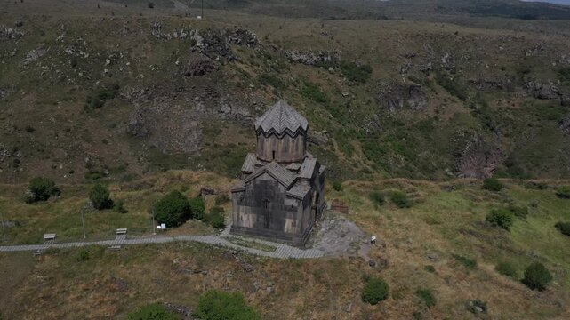 An aerial shot captures Vahramashen Church perched on a grassy hill, overlooking a dramatic gorge with mountains and a cloudy blue sky in the background.

