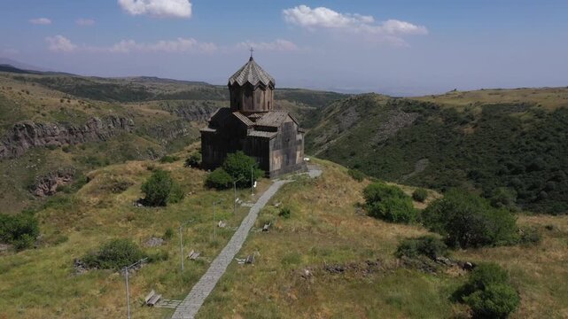 An aerial shot captures Vahramashen Church perched on a grassy hill, overlooking a dramatic gorge with mountains and a cloudy blue sky in the background.

