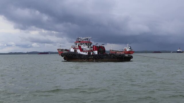 A commercial red and white tugboat vessel floating on the sea water surface against a background of dark overcast clouds and distant ships.