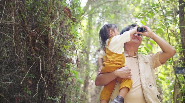 Asian father teaching young daughter using binoculars while learning about nature during travel tropical forest exploration. Parent and child enjoy outdoor adventure active lifestyle in woodland