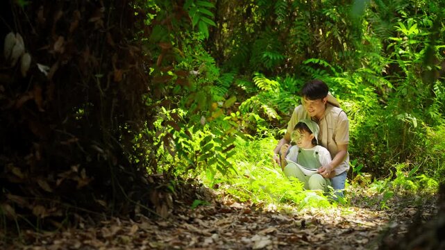 Asian father teaching daughter drawing nature journal in tropical forest, child learning about plants with magnifying glass during family hiking adventure, outdoor education and eco tourism activity.