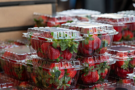 Clear plastic containers of fresh red, ripe strawberries with the stems still on them. The packages are stacked on top of each other on a grocery store shelf.  The berries are firm, rich, and vibrant.