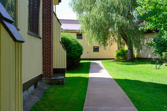 A concrete pathway among lush green manicured grass, tall mature trees, shrubs, and bushes on the side of a large religious building. The wooden structure is painted yellow with brown trim and has lar