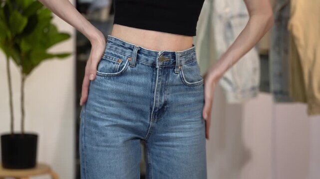 Slender woman zips and buttons high waist blue jeans, smoothing hips and turning in home interior. Concept of flattering denim fit, slim figure, body confidence, style and fashion fitting
