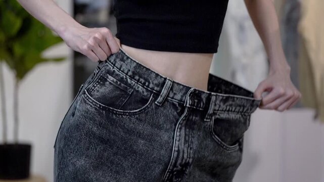 Slim woman joyfully sways hips while stretching big jeans sideways after weight loss, home interior. Concept of healthy lifestyle, diet success, wellness progress, body transformation and motivation